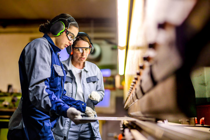 Two focused Caucasian women in safety gear use a press brake machine, working diligently in a well-lit manufacturing facility.